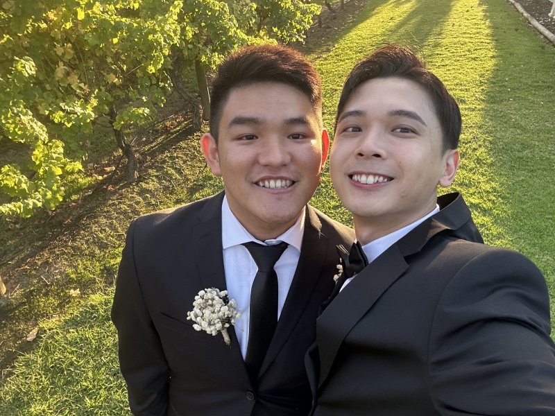 Two people in dark suits taking a selfie outdoors beside green vines.
